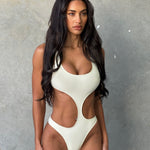 Model wearing an ivory asymmetric cut-out backless swimsuit with a high waistline, standing in front of a grey wall.