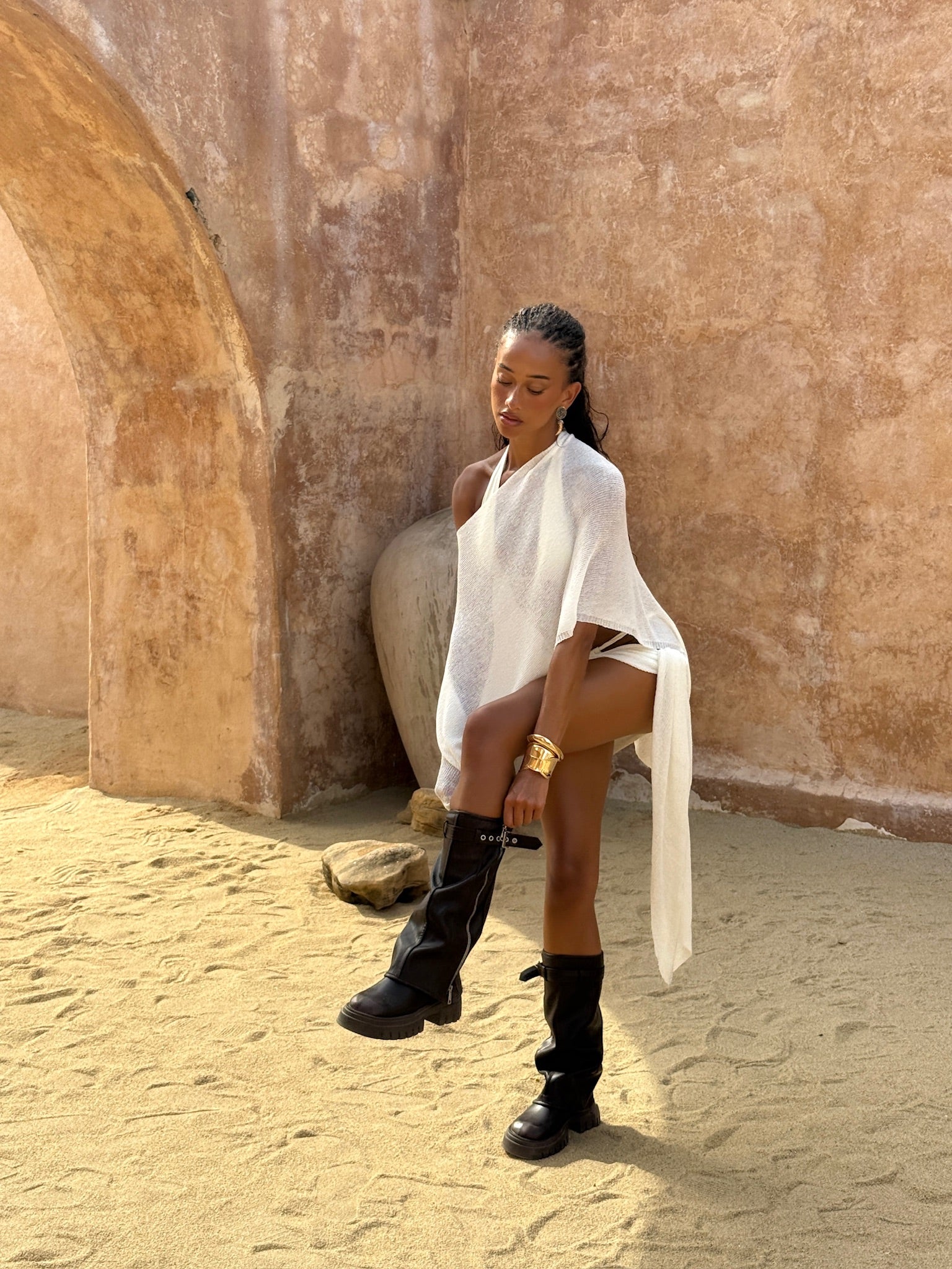 Model wearing an ivory bikini and matching ivory cape and sarong styled back with black boots sitting against a textured wall.