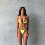 Close-up of model wearing a yellow triangle bikini top with blue trim and Rogue Season logo, paired with a matching yellow high-cut bikini bottom with blue trim, against a neutral grey background.