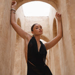 Model wearing a black halter neck maxi dress standing in a sunlit archway with sand-coloured walls.