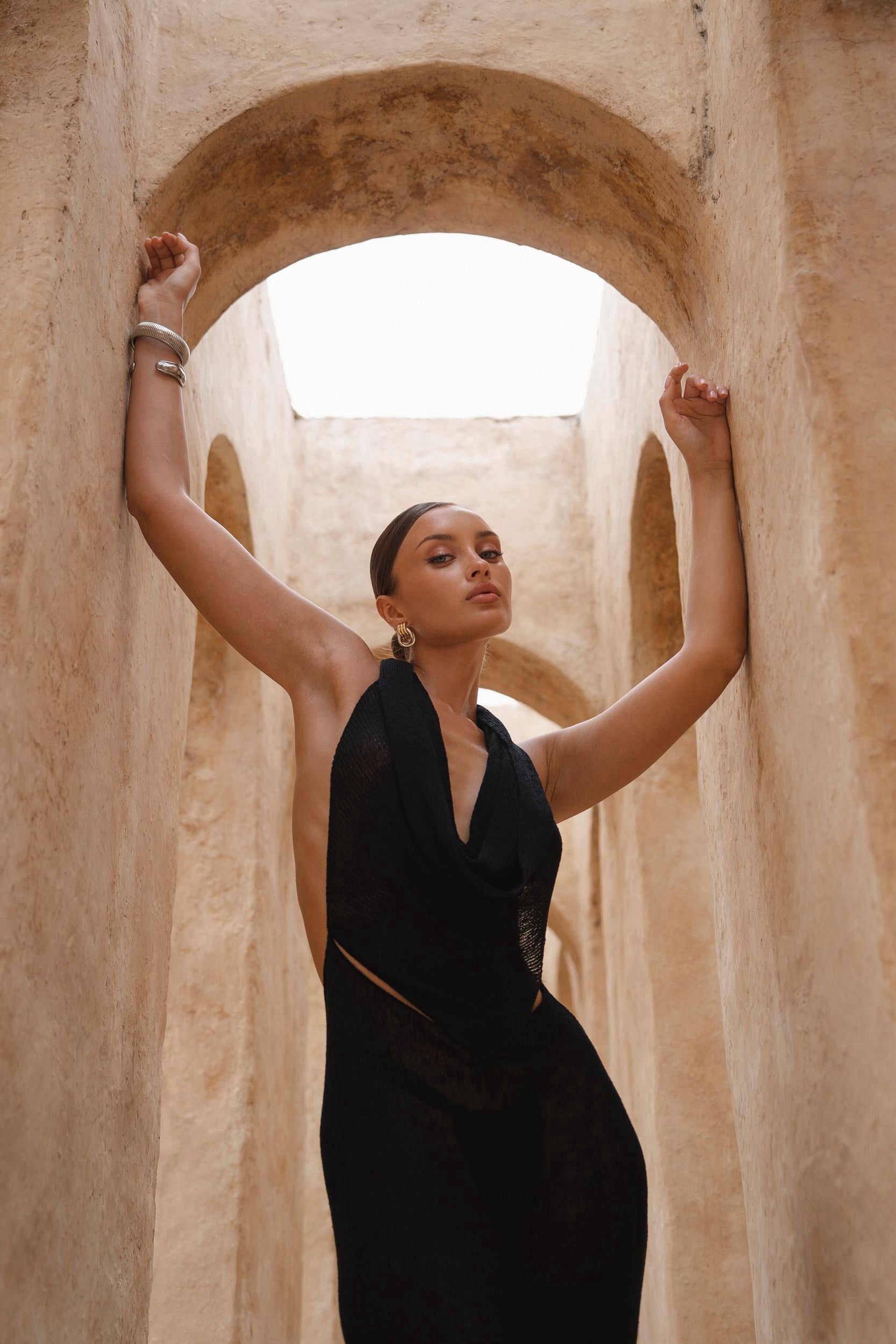Model wearing a black halter neck maxi dress standing in a sunlit archway with sand-coloured walls.