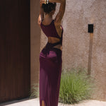 Back shot of model in a purple swimsuit standing outdoor in sunlight against a textured wall.