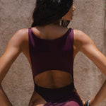 Close up back shot of model wearing a burgundy swimsuit and matching sheer maxi skirt near a textured wall