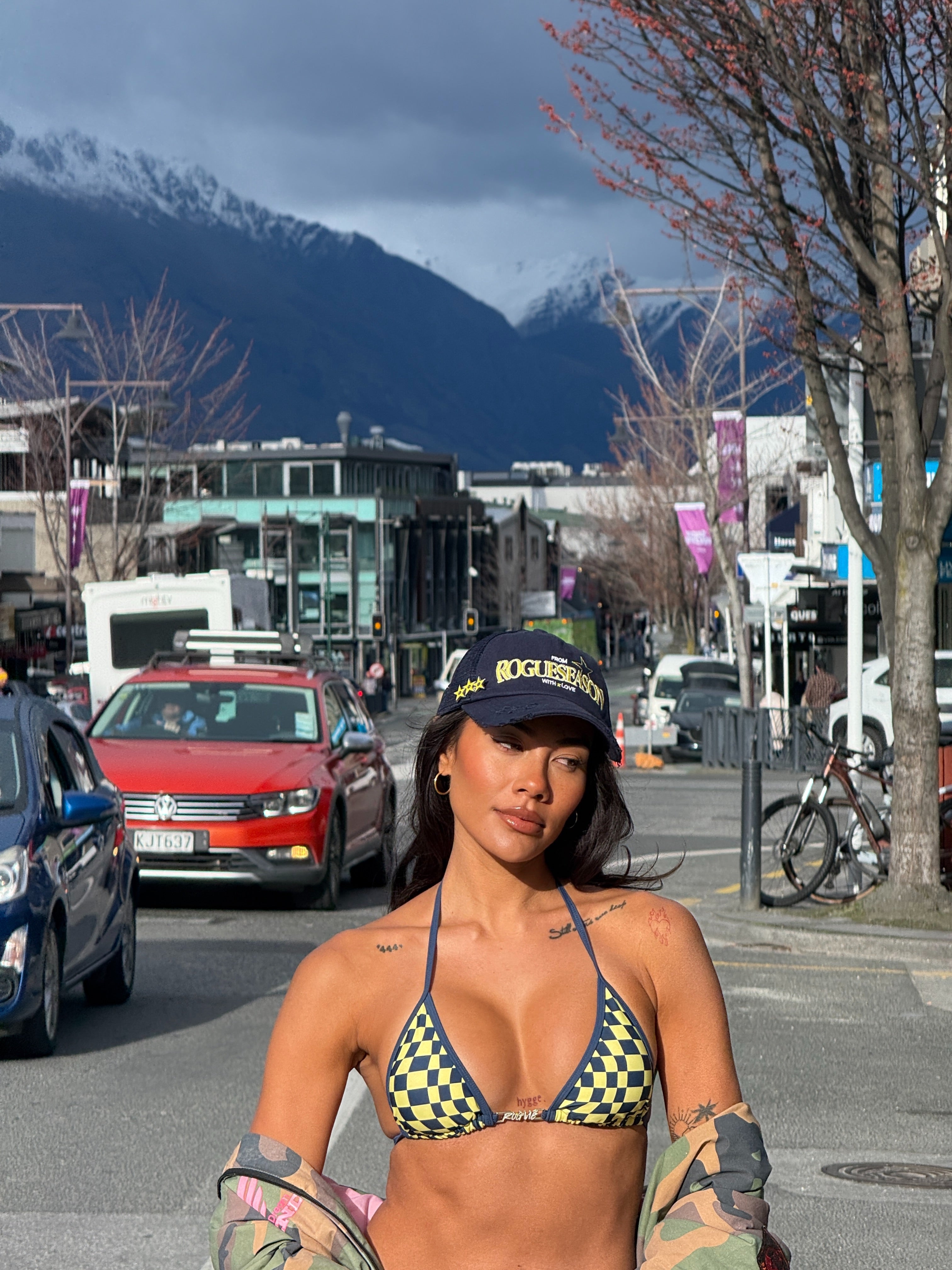 Model wearing a checkered bikini in Navy and Yellow and 'ROGUESEASON' branded cap standing on a street with mountains in the background