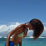 Model wearing a  green triangle bikini with navy trim and RS logo detailing, standing on sand flicking hair at the beach with sunlit skin and water droplets.