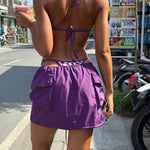 Model wearing a purple checkered bikini and purple cargo skirt on a street with shops and signs in the background