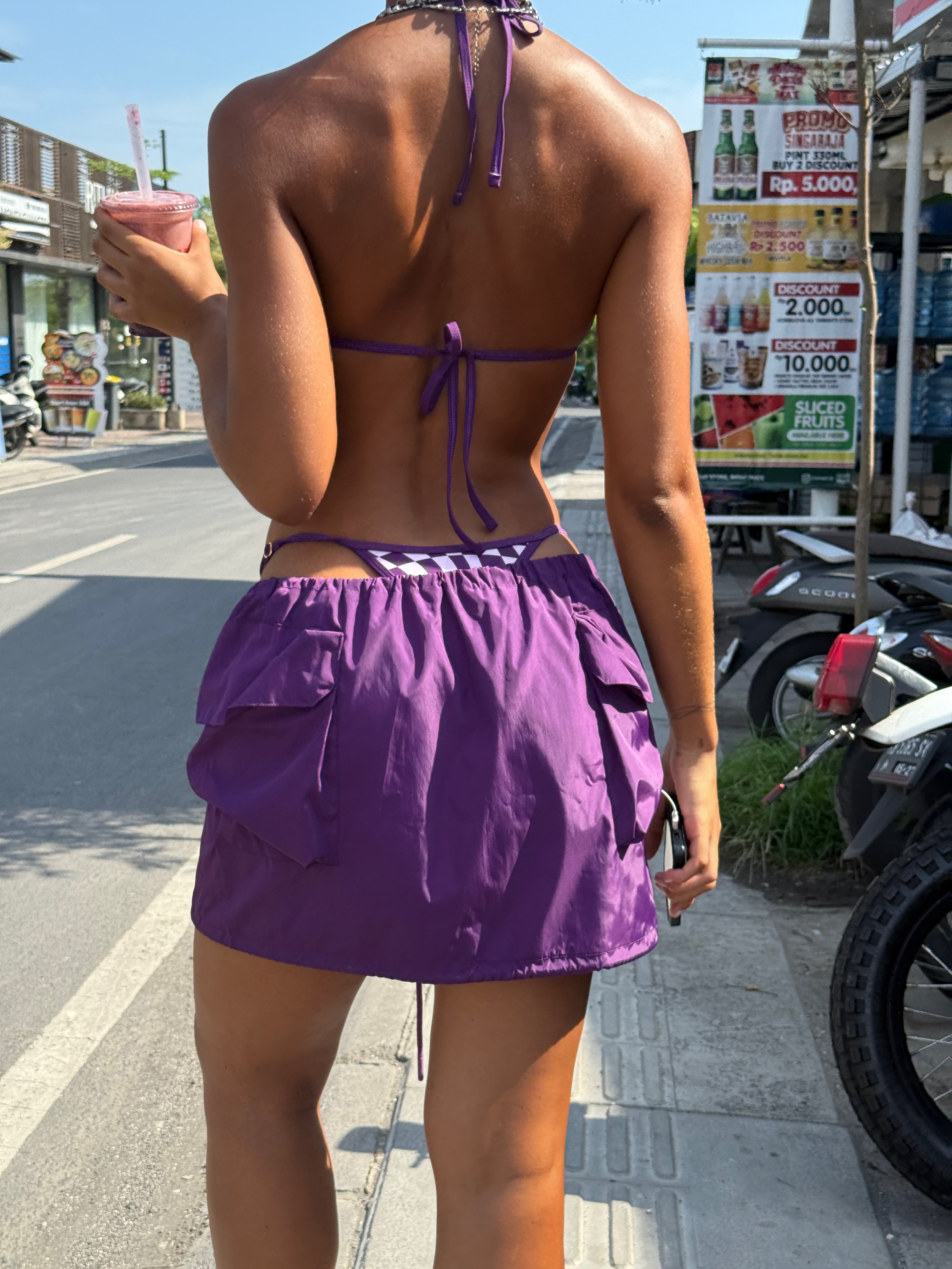 Model wearing a purple checkered bikini and purple cargo skirt on a street with shops and signs in the background