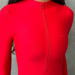 Close up model wearing a red long sleeve zip up one piece swimsuit standing next to a grey wall.