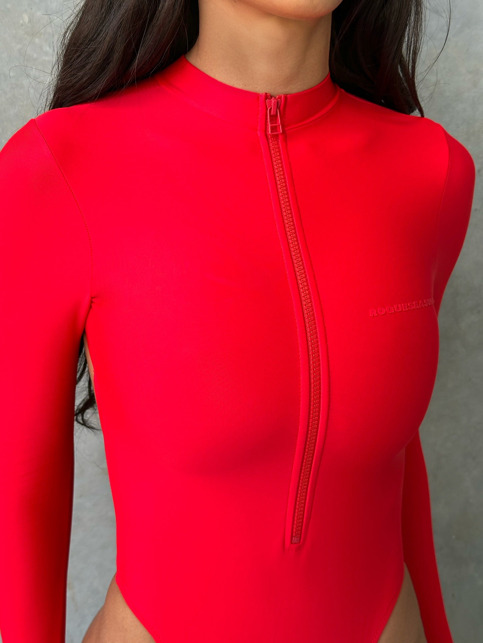 Close up model wearing a red long sleeve zip up one piece swimsuit standing next to a grey wall.