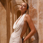 Close up side angle shot of model wearing an Ivory halter neck maxi dress standing in a sunlit archway with sand-coloured walls.