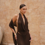 Model wearing a brown halter neck maxi dress standing in a sunlit setting with sand-coloured walls.
