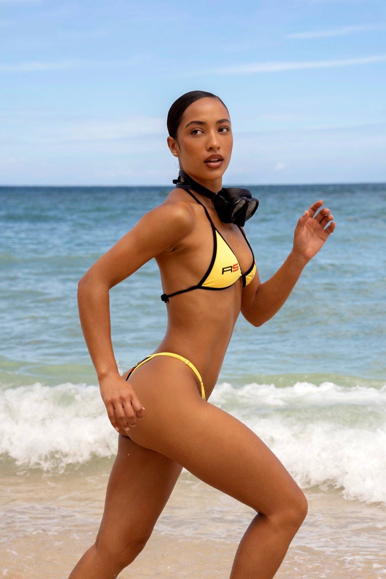 Model in Rogue Season yellow and black bikini running on a beach with ocean waves in the background