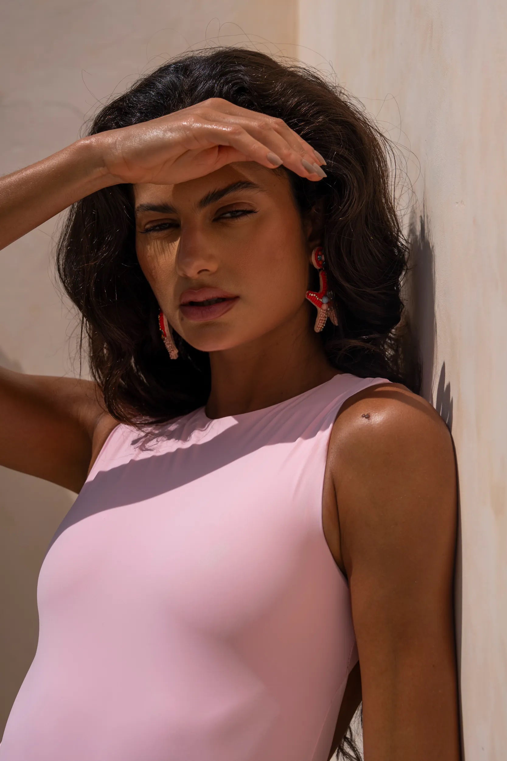 Close up shot of model covering her face from the sun while wearing a pink swimsuit and statement earrings.