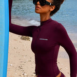 Model wearing a long-sleeve burgundy full-zip swimsuit and black sunglasses stands on a sandy beach with water in the background.