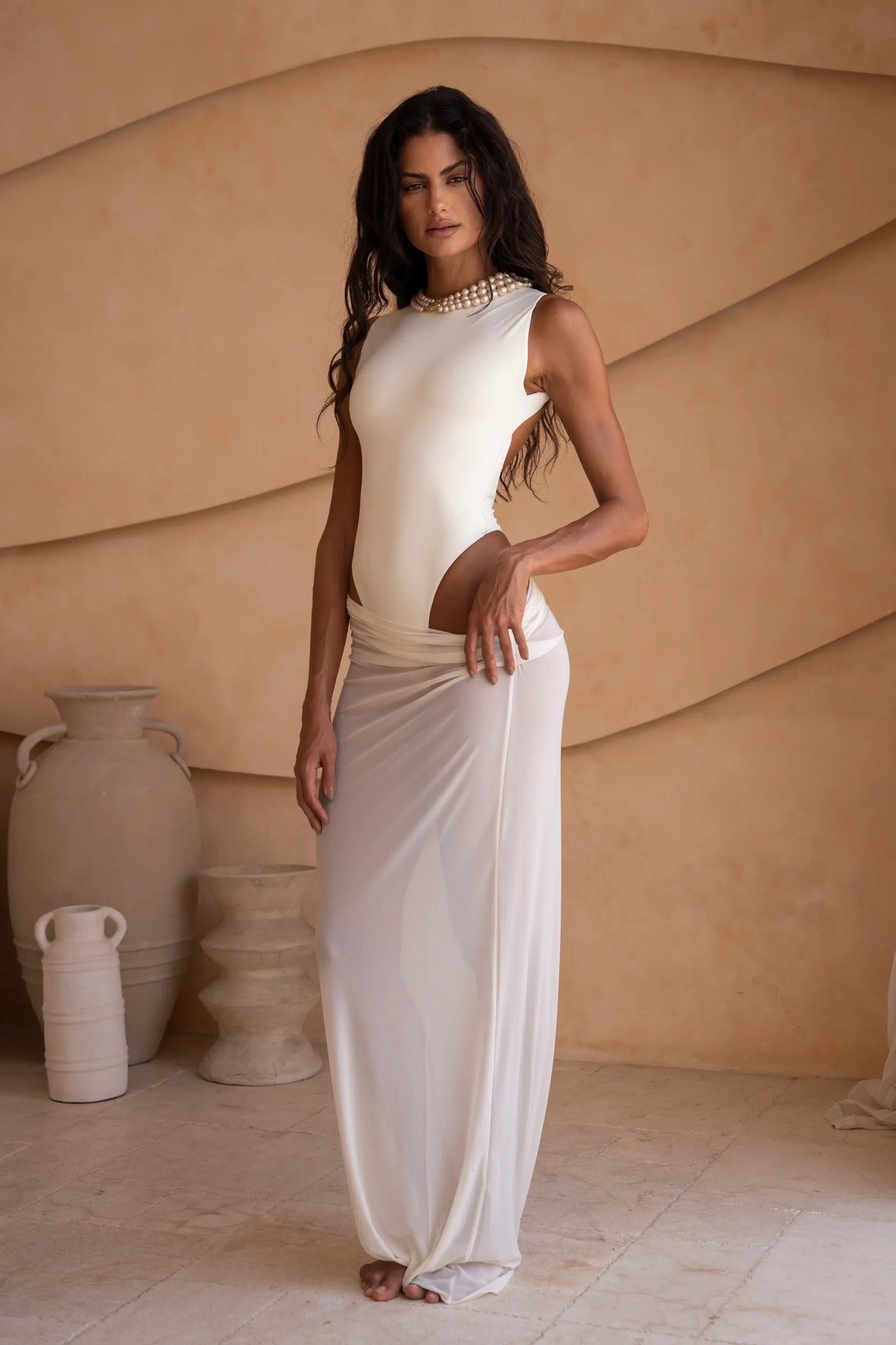 Side shot of model wearing an ivory bodysuit with a matching sheer maxi skirt with a fitted silhouette, standing indoors against a neutral-toned wall with minimal decor.