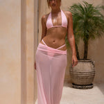 Model wearing a soft blush pink triangle bikini with adjustable halter straps and minimal-coverage bottoms with a matching sheer maxi skirt. Photographed outdoors, the image highlights the bikini's clean lines, flattering fit and elevated resort-inspired design.