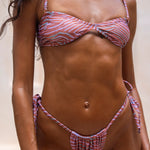 Crop in shot of model wearing a coral print bikini set featuring a bandeau-style top with thin straps and tie-side Brazilian bottoms. The model stands against a warm-toned textured backdrop, showcasing the swimwear's flattering fit, minimal coverage and adjustable side ties.