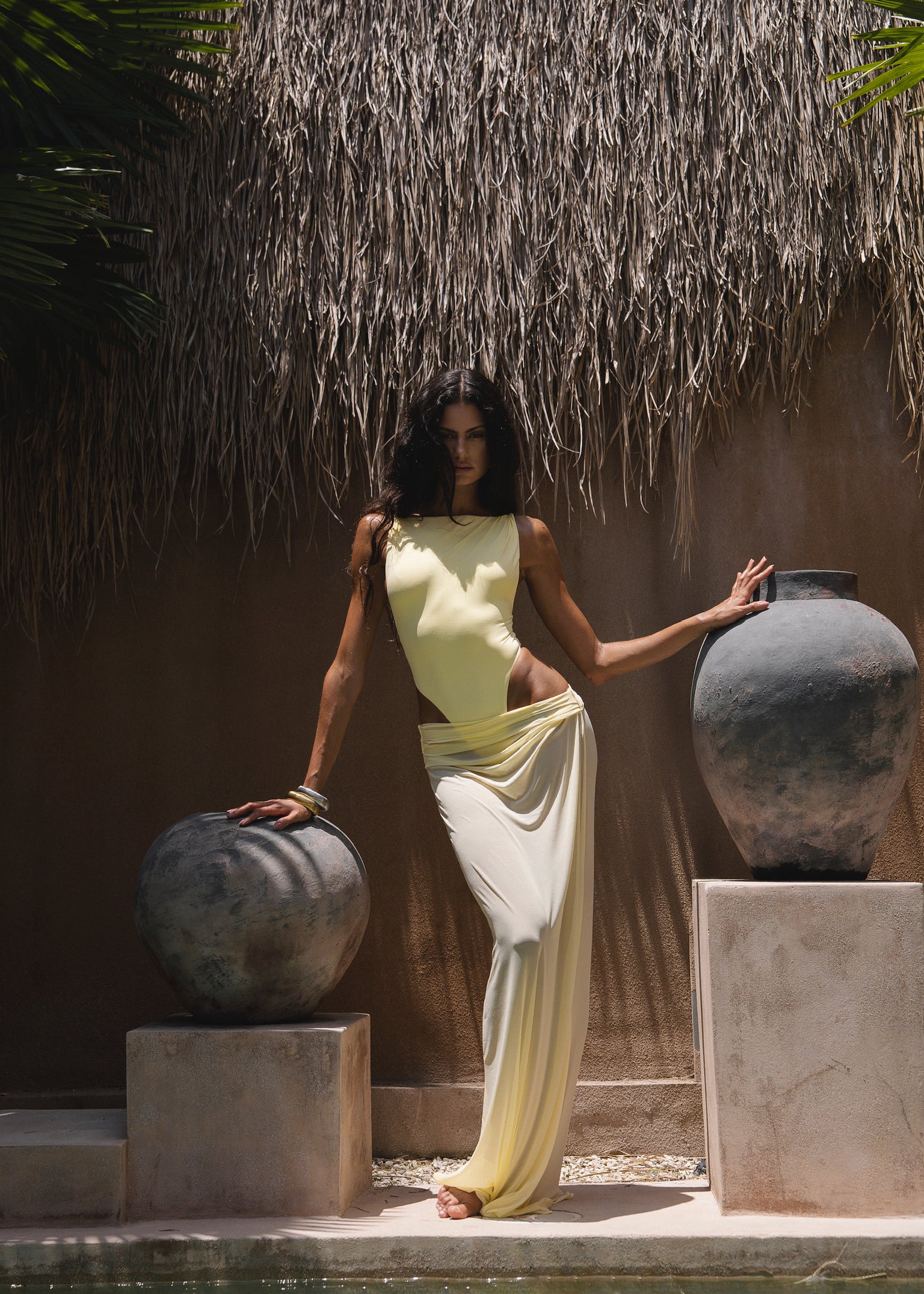 Model in a yellow swimsuit with a matching sheer maxi skirt, standing between two large pots in a tropical setting