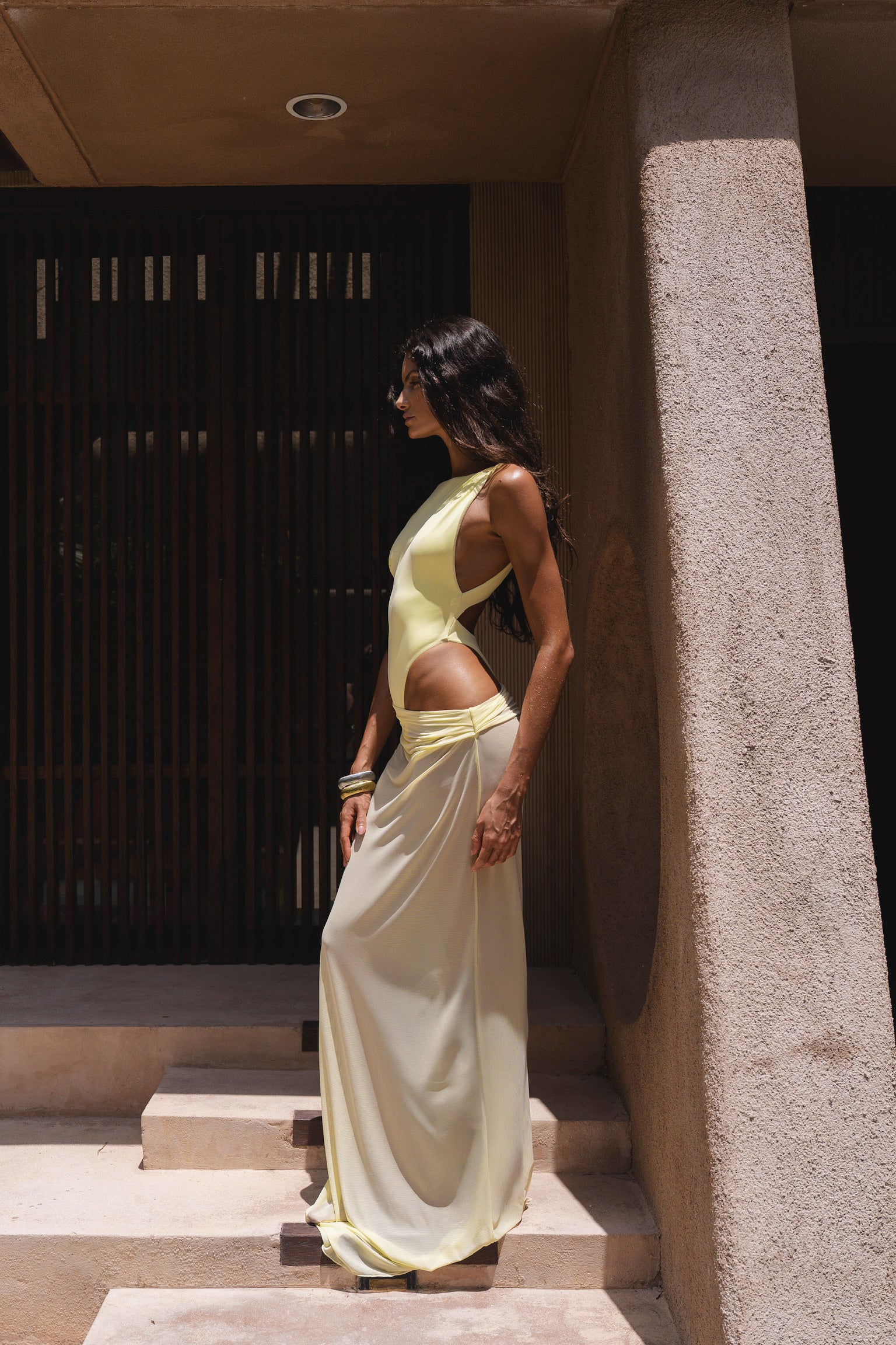 Model in a yellow swimsuit and matching sheer maxi skirt standing on steps with a building in the background