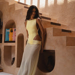 Model in a yellow swimsuit and matching sheer maxi skirt, standing against a textured staircase and wall with geometric shapes.
