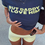 Model wearing a navy blue t-shirt with yellow text 'BUT DADDY I LOVE HIM' and light yellow shorts.