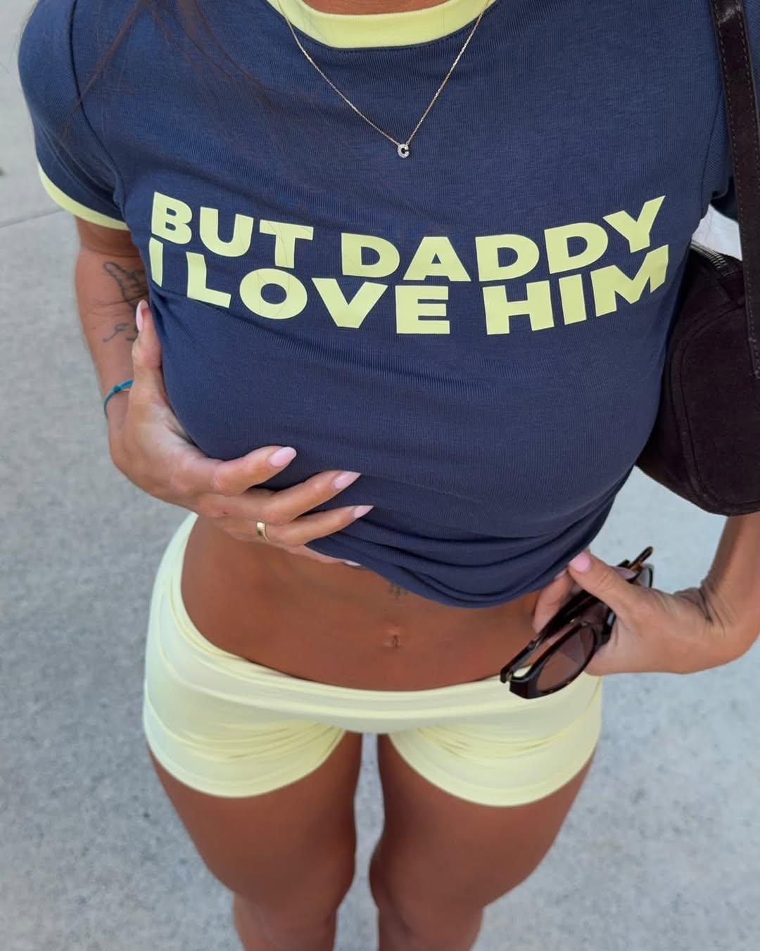 Model wearing a navy blue t-shirt with yellow text 'BUT DADDY I LOVE HIM' and light yellow shorts.